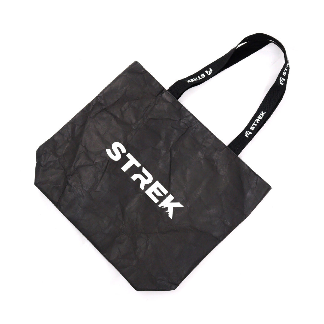 Flat lay angled view of the STREK Overland Tyvek Utility Tote Bag showing the black Tyvek body and branded carry straps.