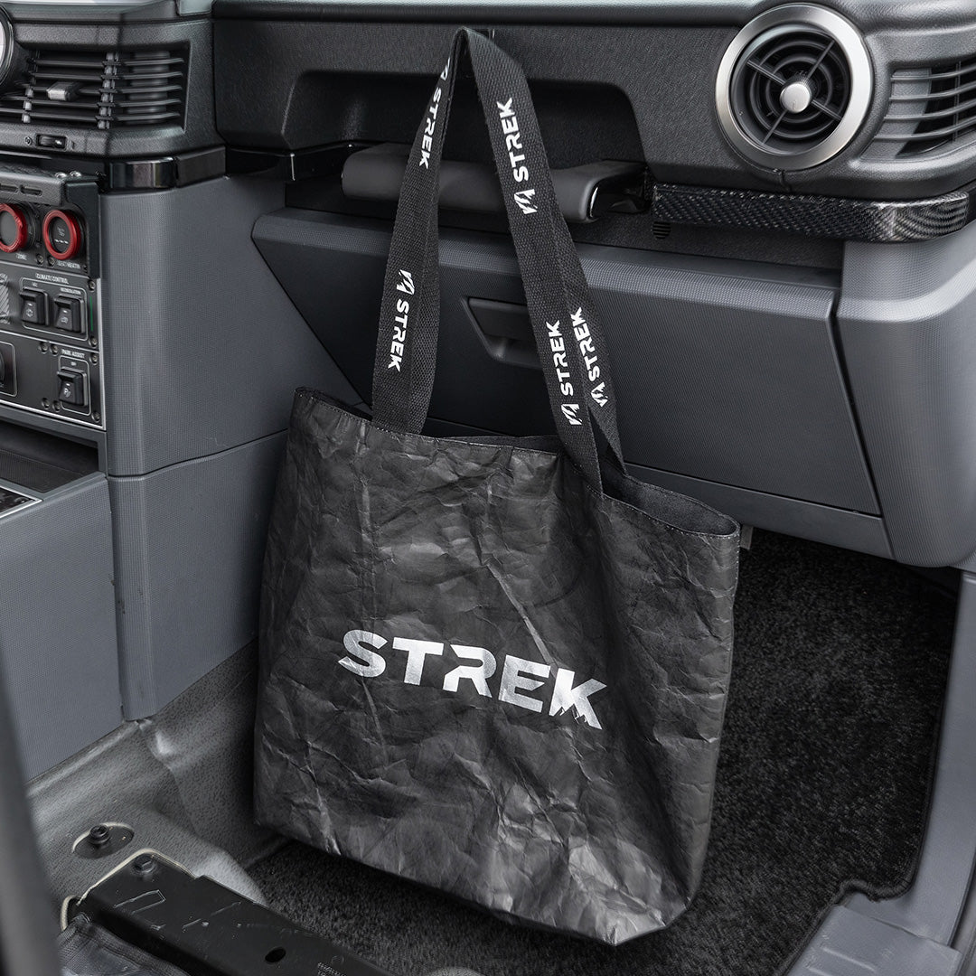 STREK Overland Tyvek Utility Tote Bag placed in an INEOS Grenadier passenger footwell with branded handles looped near the dashboard grab handle.