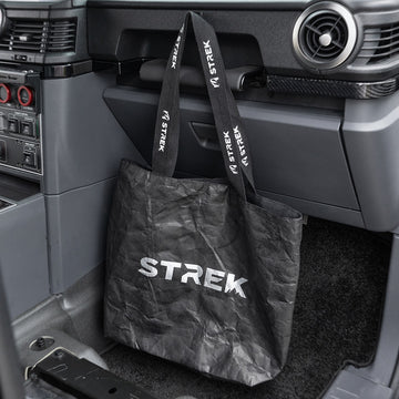 STREK Overland Tyvek Utility Tote Bag placed in an INEOS Grenadier passenger footwell with branded handles looped near the dashboard grab handle.